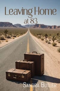 The front cover of "Leaving Home at 83." Depicts a long highway in the desert disappearing into the horizon and hills. In the middle of the road are three old brown suitcases.