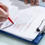 A photograph of a hand with a sheets of paper on a clipboard, using a red pen to make edits to the document.