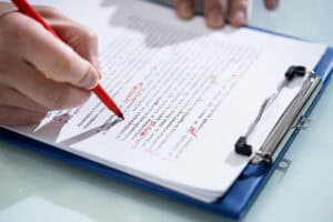 A photograph of a hand with a sheets of paper on a clipboard, using a red pen to make edits to the document.