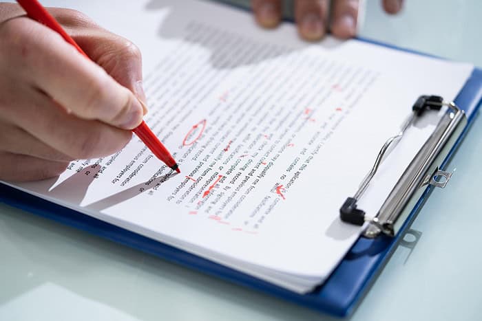 A photograph of a hand with a sheets of paper on a clipboard, using a red pen to make edits to the document.
