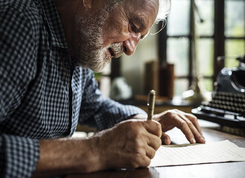 Close-up of an older man smiling softly as he writes by hand at a wooden desk, absorbed in his work.