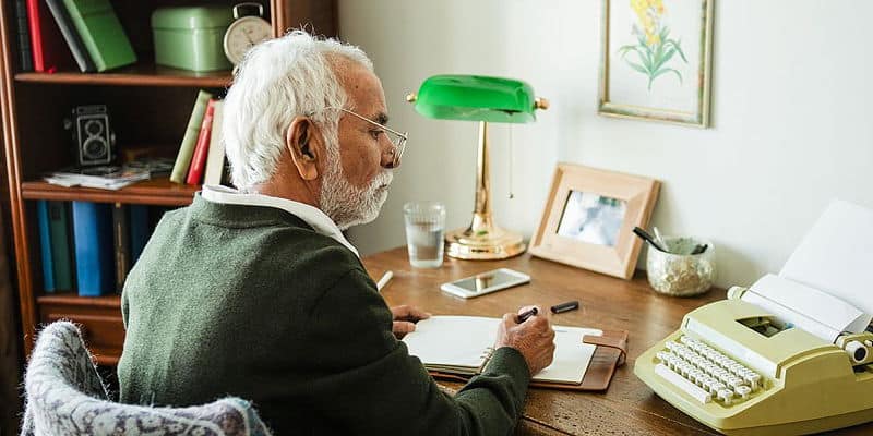 Older man sitting at a home desk with a notebook and vintage typewriter, focused on writing in a cozy, book-lined room.