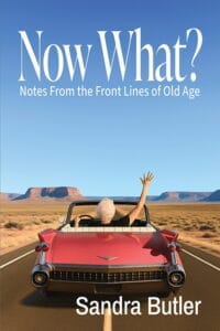 Cover of Now What? Notes From the Front Lines of Old Age by Sandra Butler. A white-haired older woman drives a red vintage convertible down a long desert highway, raising one hand in the air, with mesas in the distance beneath a wide blue sky.